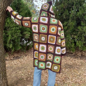 May include: A hooded, long-sleeved crochet coat in a patchwork design. The coat features squares in shades of brown, green, white, and yellow. The person is wearing blue jeans. The coat is a unique, handmade garment.