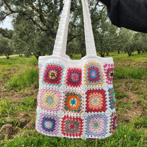 May include: White crochet tote bag with colourful square patches. The bag has a long strap and is made of a soft, textured material.