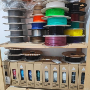 May include: A shelf with multiple spools of filament for 3D printing. The spools are arranged in rows and are of various colors, including gray, green, blue, red, and yellow. The spools are labeled with the brand name "SMARTFIL".