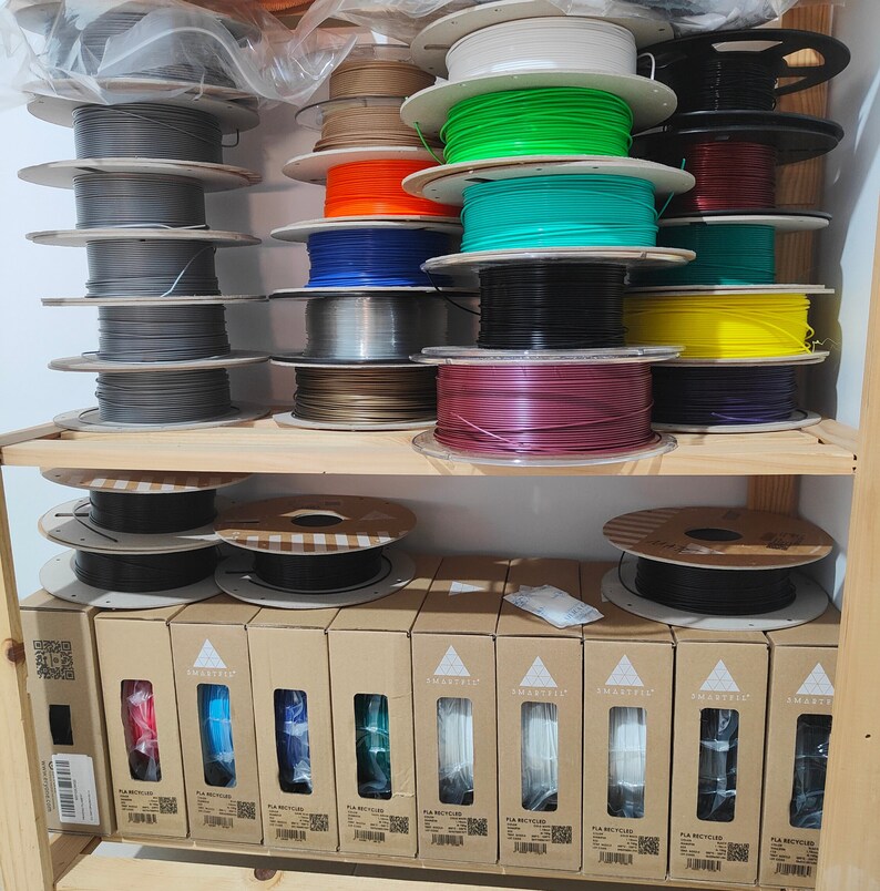 May include: A shelf with multiple spools of filament for 3D printing. The spools are arranged in rows and are of various colors, including gray, green, blue, red, and yellow. The spools are labeled with the brand name "SMARTFIL".