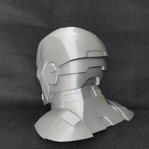 May include: A gray 3D printed helmet with a detailed design, resembling a futuristic or sci-fi character. The helmet has a visor and a neck piece.