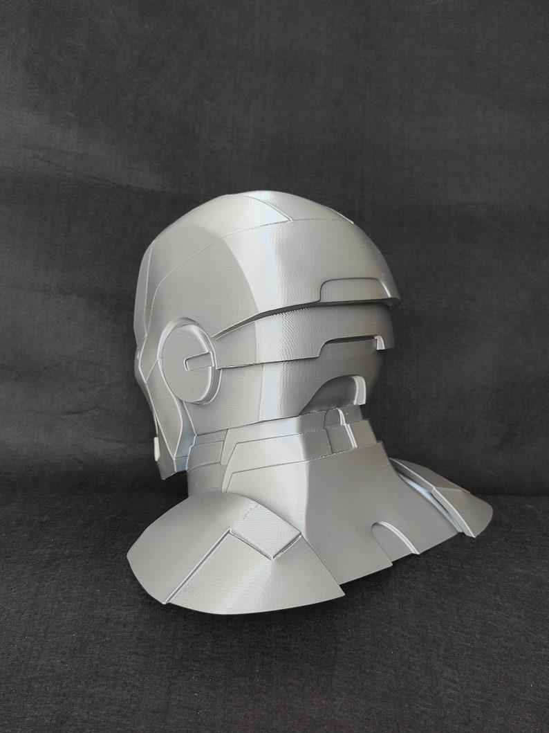 May include: A gray 3D printed helmet with a detailed design, resembling a futuristic or sci-fi character. The helmet has a visor and a neck piece.
