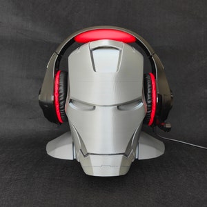 May include: A gray 3D printed Iron Man head with a black headset with red ear cushions. The headset is resting on the Iron Man head.