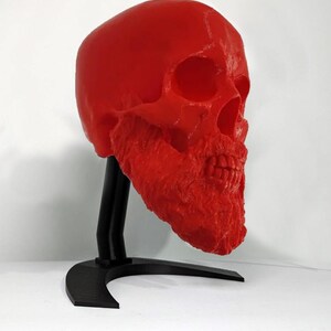 May include: A vibrant red skull sculpture with a textured surface, mounted on a black stand. The skull has detailed features and a prominent jawline. The stand has a curved base and a vertical support.