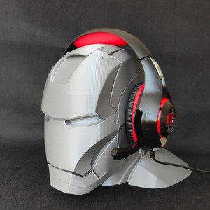 May include: A black and red gaming headset with a microphone is displayed on a gray 3D printed stand shaped like a helmet.