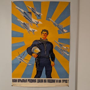 Pode incluir: Cartaz vintage com um piloto em uniforme azul escuro segurando um capacete, com caças contra um fundo de raios de sol amarelo e azul. O cartaz tem texto em russo na parte inferior.
