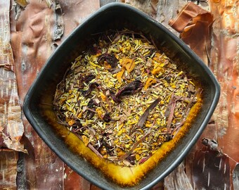 May include: A black ceramic bowl filled with a blend of dried herbs and flowers. The bowl is sitting on a background of brown and orange bark.