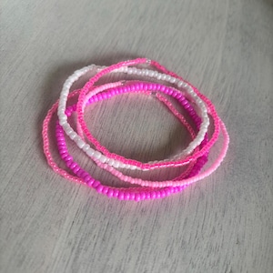 May include: A set of beaded bracelets in various shades of pink and white. The bracelets are composed of small, round beads, forming a delicate accessory. The colours vary from pale pink to a bright fuchsia.