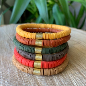 Puede incluir: Pila de seis pulseras de cuentas de colores. Las pulseras presentan una variedad de colores tierra, incluyendo amarillo, marrón, verde, gris, coral y beige. Cada pulsera tiene un acento metálico dorado. Las pulseras están apiladas sobre una superficie de madera.