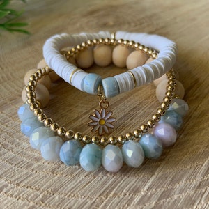 May include: A set of three bracelets featuring a white beaded bracelet, a wooden beaded bracelet, and a gold beaded bracelet with light blue and pink faceted beads. A gold charm with a white daisy is attached to the gold beaded bracelet.