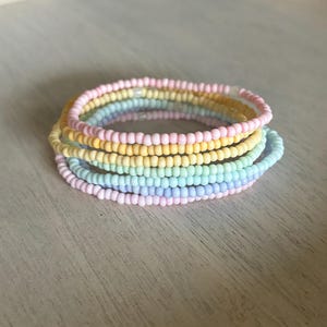 Może przedstawiać: Stos bransoletek z koralików w pastelowych kolorach. Bransoletki są w odcieniach różu, żółci, zieleni i błękitu. Bransoletki są ułożone jedna na drugiej, tworząc efekt tęczy. Koraliki są małe i okrągłe.