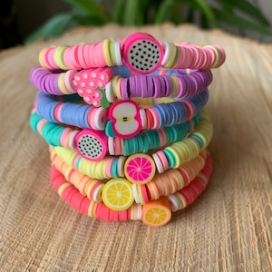 Op de afbeelding: Een stapel kleurrijke armbanden met fruitbedels. De armbanden zijn gemaakt van roze, paarse, blauwe, gele en oranje kralen. De bedels zijn een watermeloen, een tros druiven, een appel, een grapefruit en een citroen.