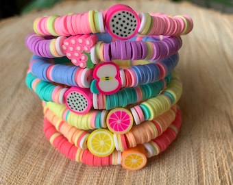 May include: A stack of colorful bracelets with fruit charms. The bracelets are made of pink, purple, blue, yellow, and orange beads. The charms include a watermelon, a bunch of grapes, an apple, a grapefruit, and a lemon.