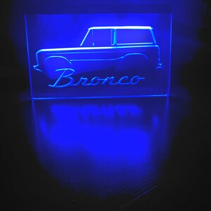 May include: A blue illuminated sign with a silhouette of a Ford Bronco and the word "Bronco" underneath.