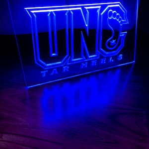 Könnte beinhalten: Blaues Neon-Schild mit dem Wort "UNC" und den Worten "Tar Heels" darunter. Das "C" in UNC ist ein stilisierter Fußabdruck.