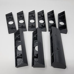 May include: A collection of black, 3D-printed plastic components. The pieces have a geometric design with various cutouts and holes. The parts are arranged on a white surface, showcasing their shape and structure. The components appear to be for a larger assembly.