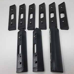 May include: A collection of black door hardware components, including strike plates and latch guards, arranged against a white background. The pieces have rectangular and elongated shapes with various cutouts and screw holes. The hardware is likely for door security.