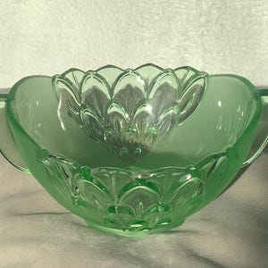 May include: A light green glass bowl with a decorative floral design. The bowl has two handles and a smaller, matching bowl inside. The glass has a smooth, glossy finish.