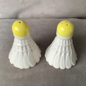 May include: A pair of white ceramic salt and pepper shakers shaped like badminton shuttlecocks with yellow tops.