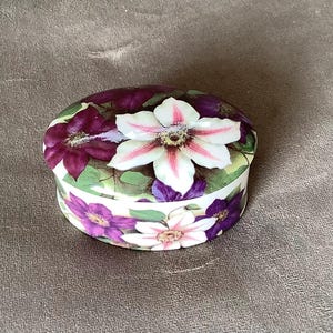 May include: Oval ceramic trinket box with a floral design. The lid features a large white and pink flower surrounded by purple flowers and green leaves. The sides of the box are decorated with smaller purple and white flowers.