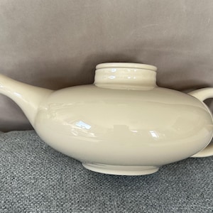 May include: Cream-colored ceramic teapot with a rounded body, a curved spout, and a loop handle. The lid sits atop the teapot, and the piece has a glossy finish. The teapot is designed for serving tea.