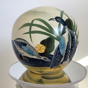 以下が含まれることがあります： 丸い形の白い陶器の花瓶で、2羽の鳥と緑の葉の模様が描かれています。鳥は青、黒、白で、黄色いアクセントが付けられています。