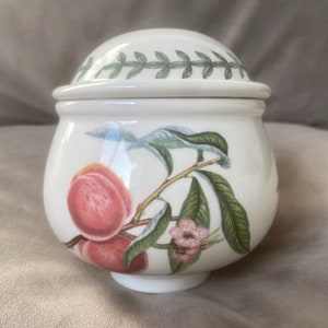 Può includere: Zuccheriera in ceramica bianca con coperchio, con un motivo di pesca con foglie verdi e fiori rosa. Il coperchio ha un bordo di foglie verdi.