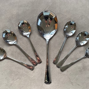 May include: A set of seven silver spoons with a decorative fan-shaped design on the bowl. The spoons are arranged in a circle on a gray surface.