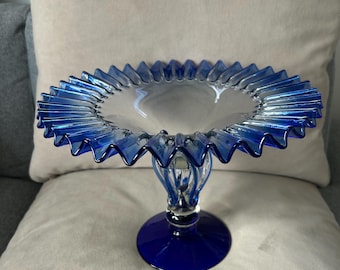 May include: A clear glass pedestal bowl with a vibrant blue ruffled rim and base. The bowl has a clear, circular center and a decorative, fluted stem. The base is a solid, glossy blue. This elegant piece is suitable for serving or display.