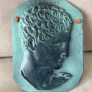 May include: A rectangular teal plaque featuring a dark relief profile of a person with curly hair. The text OLYMPIE is embossed at the top, and HERMES is embossed vertically on the right side. Two small orange cords are threaded through holes.
