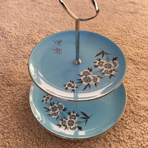 May include: Two-tiered cake stand with a silver handle. The stand is decorated with a blue background and white flowers.