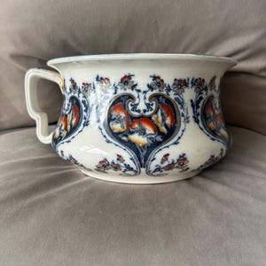 May include: Antique white porcelain chamber pot with a handle. The pot features a decorative floral design in shades of blue, orange, and red. The rim and handle are white.