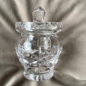 Puede incluir: Un tarro de vidrio transparente con un diseño de cristal tallado y una tapa a juego. El tarro tiene un cuerpo redondo y una boca ancha.