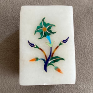 May include: A white marble box with a floral design inlaid with colorful stones. The flower is green with blue and orange accents. The box is a decorative piece.