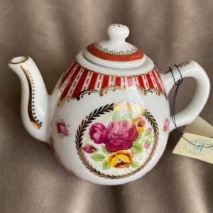 Könnte beinhalten: Eine weiße Porzellan-Teekanne mit rotem und goldenem Blumendesign. Die Teekanne hat einen Deckel mit einem kleinen Knopf oben. Die Teekanne ist mit einem roten und goldenen Streifenband um die Mitte verziert. Die Teekanne steht auf einer beigefarbenen Oberfläche.