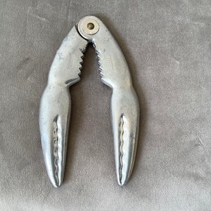 Puede incluir: Cascanueces de cangrejo de plata con una superficie texturizada y un pequeño pomo de metal en la parte superior. El cascanueces está diseñado para parecer una pinza de cangrejo.