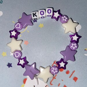Puede incluir: Un brazalete de cuentas con forma de estrella de color morado y blanco con las letras "KOO" escritas en cuentas blancas.