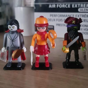 Puede incluir: Tres figuras de plástico en miniatura, un vampiro, una detective y un pirata. El vampiro lleva un traje gris, una capa roja y un sombrero negro. La detective lleva una camisa amarilla, una falda roja, una bufanda y gafas. El pirata lleva una camisa negra, una bandana roja y un sombrero negro. Las tres figuras están sobre bases negras.