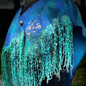 Peut inclure: Un châle en feutre bleu avec un col en V et une fermeture à bouton unique. Le châle présente un dégradé de bleus et de verts, avec des accents dorés et de longues franges bouclées dans des tons turquoise et sarcelle. Le vêtement est présenté sur un mannequin noir.