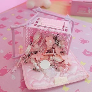 May include: A light pink miniature shopping trolley filled with pink and clear beads, flower-shaped charms, and pearls. The trolley sits on a pink quilted pad, with a pink background featuring cat and bow patterns. The trolley has the text "Nicoleskey".