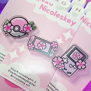 Könnte beinhalten: Drei klare Acrylstifte mit floralen Designs auf rosa Rückenkarten. Die Stifte zeigen einen Pokeball, ein Ticket und eine Handheld-Konsole, alle mit rosa Kirschblüten verziert. Die Karten tragen den Text "Nicoleskey" und Social-Media-Handles.
