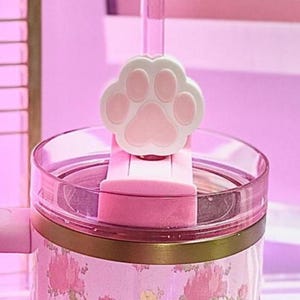 May include: Pink floral-patterned tumbler with a matching handle and lid. The tumbler has a gold band and a paw-shaped charm on the straw. The design is a cute and feminine style, ideal for cold drinks.