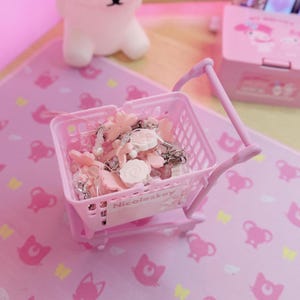 May include: A miniature pink shopping trolley filled with pink and white charms and beads. The trolley has a handle and wheels, and is on a pink surface with a repeating pattern. The text "Nicoleskey" is visible on the trolley.