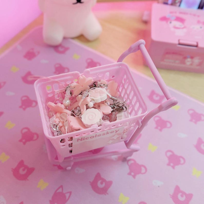 May include: A miniature pink shopping trolley filled with pink and white charms and beads. The trolley has a handle and wheels, and is on a pink surface with a repeating pattern. The text "Nicoleskey" is visible on the trolley.