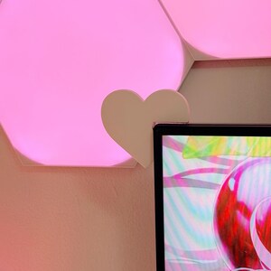 May include: A white heart-shaped clip attached to a computer monitor. The background features pink hexagonal LED lights and a pink wall. The monitor displays a colorful, abstract design.