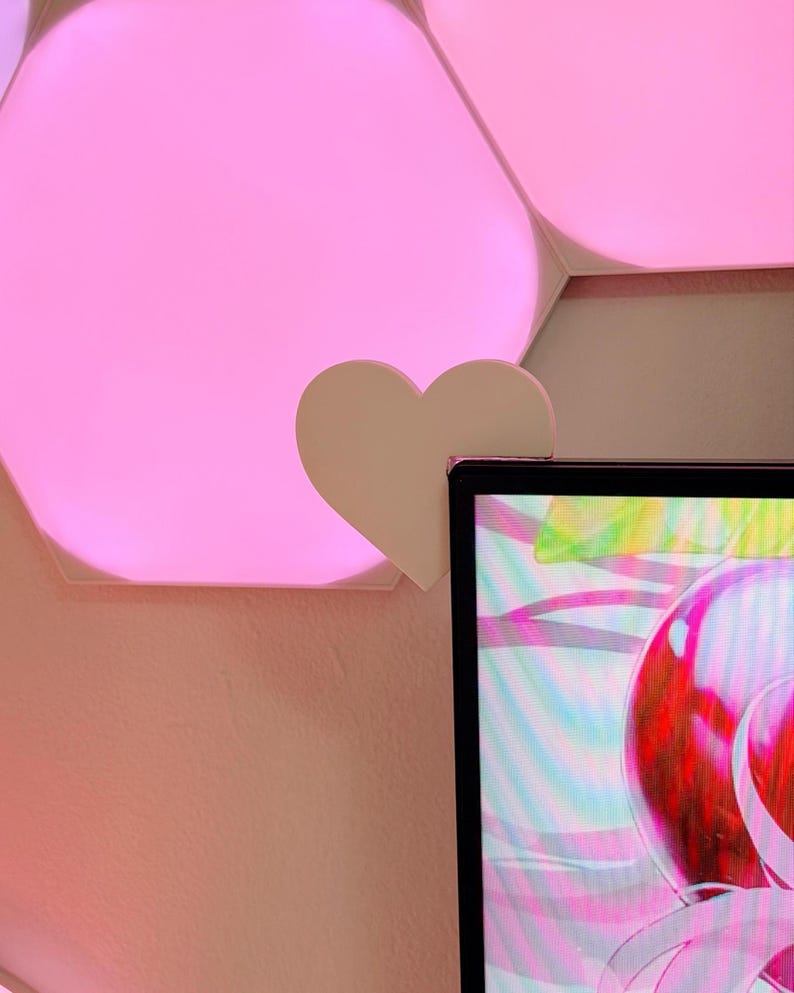 May include: A white heart-shaped clip attached to a computer monitor. The background features pink hexagonal LED lights and a pink wall. The monitor displays a colorful, abstract design.