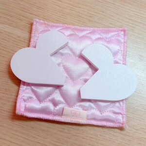 May include: Two white, heart-shaped pieces sit atop a pink, quilted square with a heart pattern. The square has a small pink ribbon with a bow design. The background is a light wooden surface.