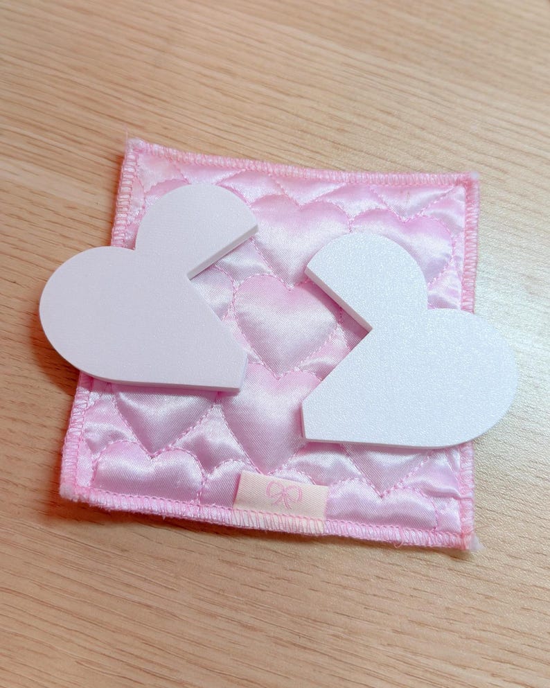 May include: Two white, heart-shaped pieces sit atop a pink, quilted square with a heart pattern. The square has a small pink ribbon with a bow design. The background is a light wooden surface.