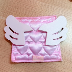 May include: A pink, quilted heart-patterned fabric square with white angel wings attached. The fabric has a satin sheen. A small pink tag is sewn on the bottom. The wings are made of a solid, smooth material.