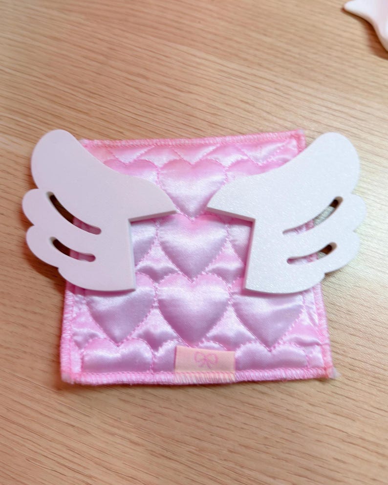 May include: A pink, quilted heart-patterned fabric square with white angel wings attached. The fabric has a satin sheen. A small pink tag is sewn on the bottom. The wings are made of a solid, smooth material.